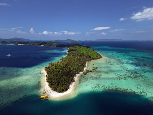 A lone forested island with a white sand tip and an outrigger boat in turquoise water, Palawan